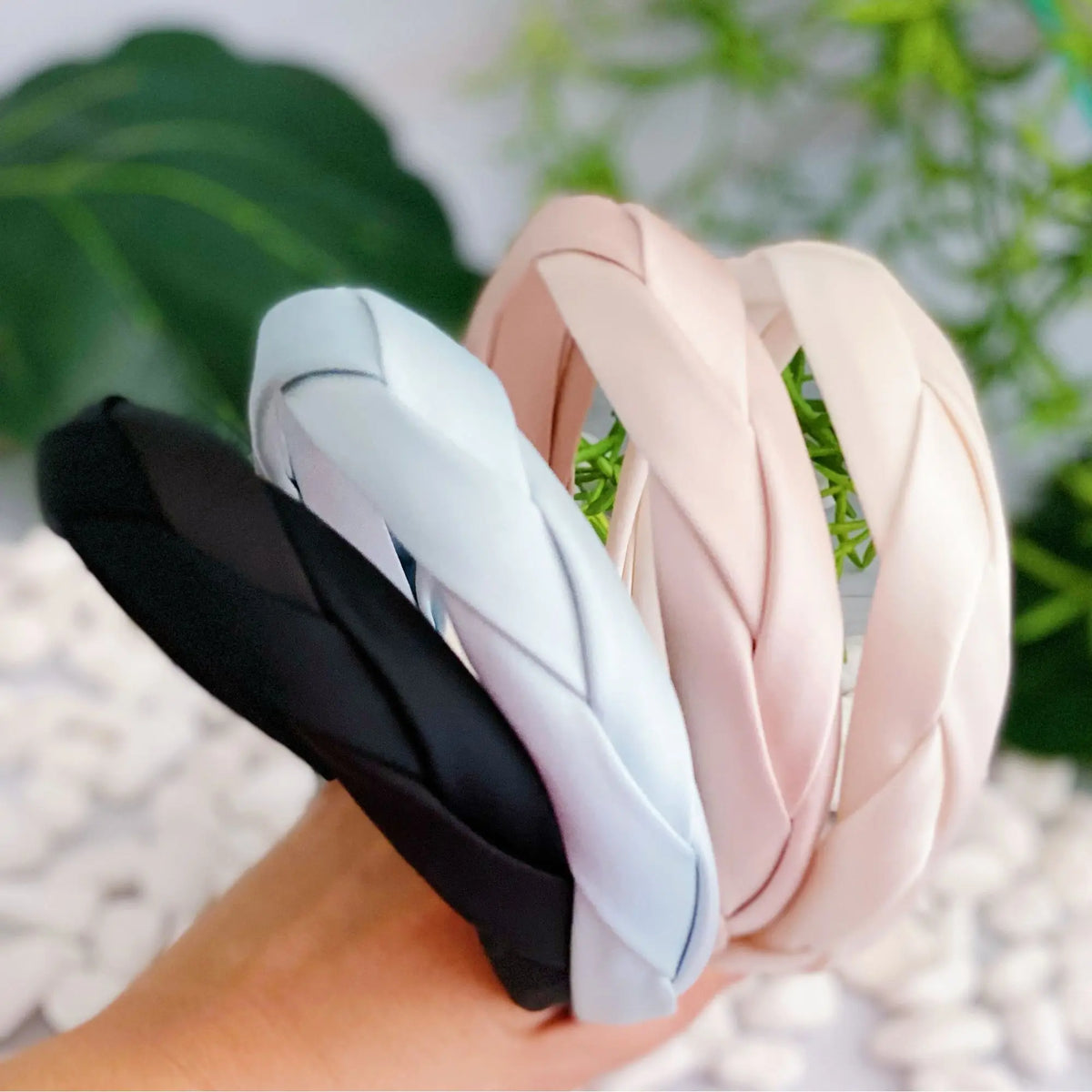 Three satin-like twisted headbands in black, light blue, and blush pink are held in a hand against a soft background with green leaves and white pebbles.